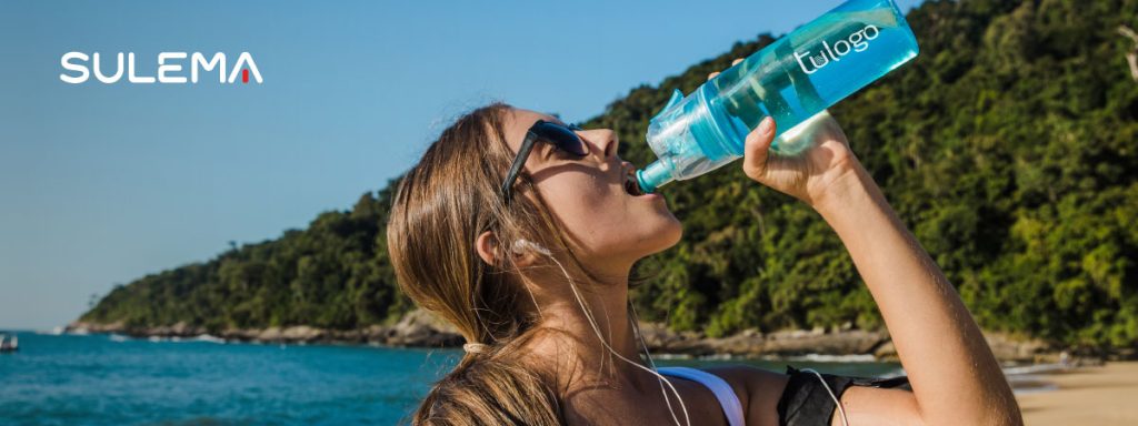 Mujer joven tomando agua de una botella reutilizable personalizada, en una playa en verano